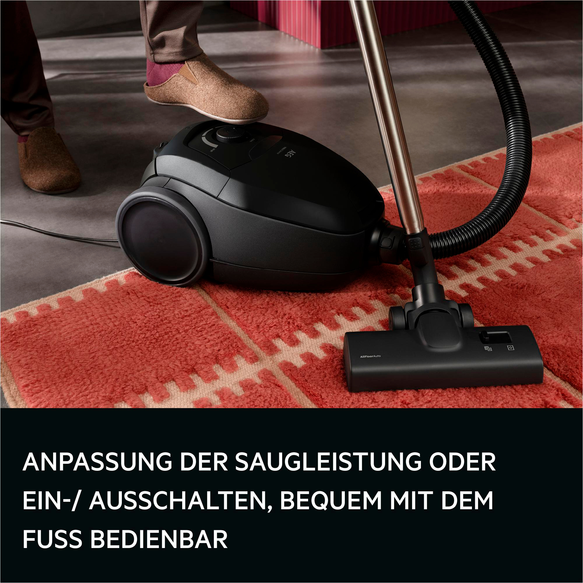 Schwarzer AEG Staubsauger mit Schlauch auf rotem Teppich, Fuß bedient ihn.