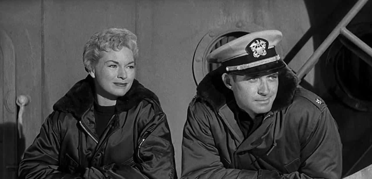 Ein Mann in Marineuniform und eine Frau in Bomberjacke sitzen eng beieinander.