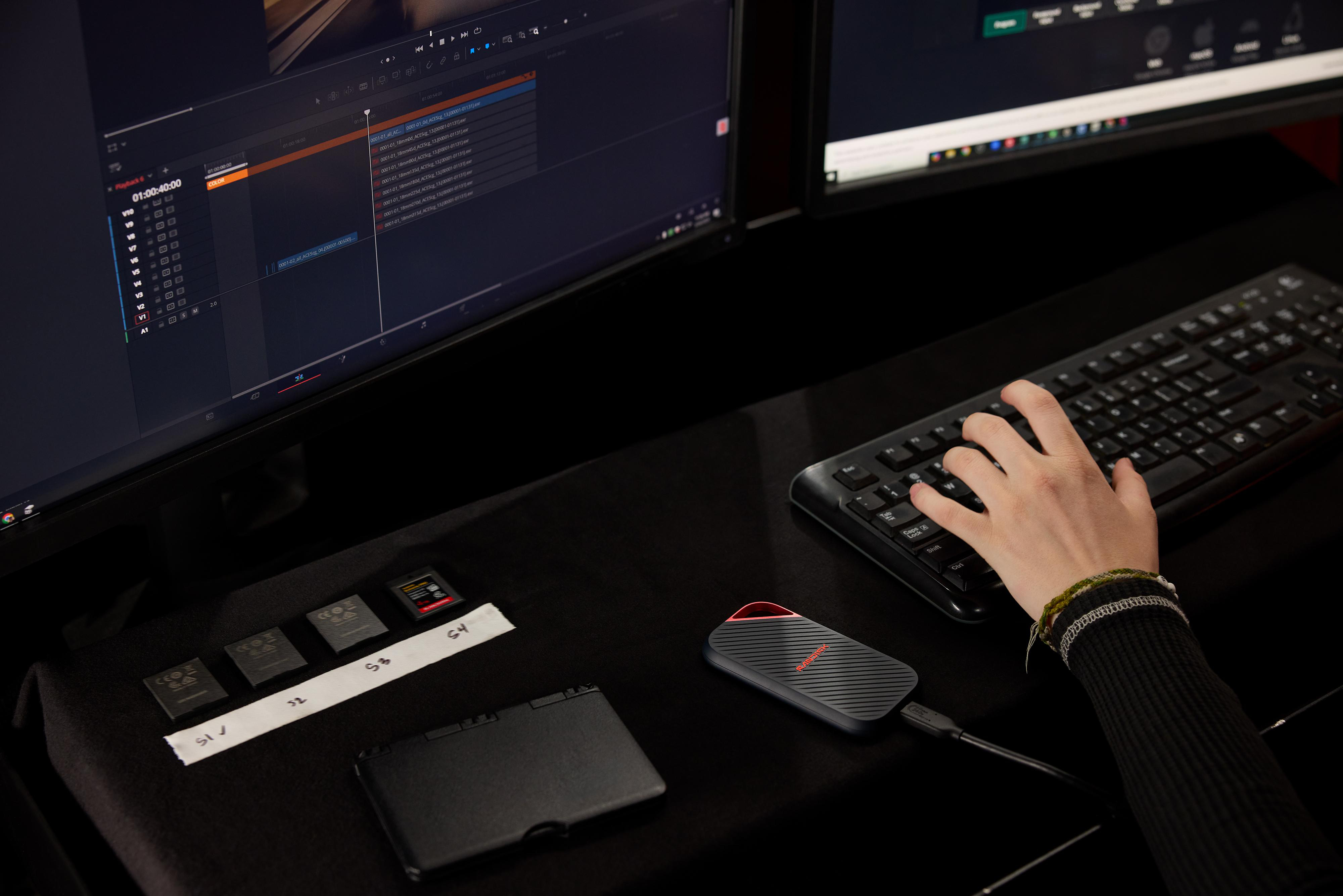 Person bearbeitet Video auf zwei Monitoren, mit externer SSD und Speicherkarten.