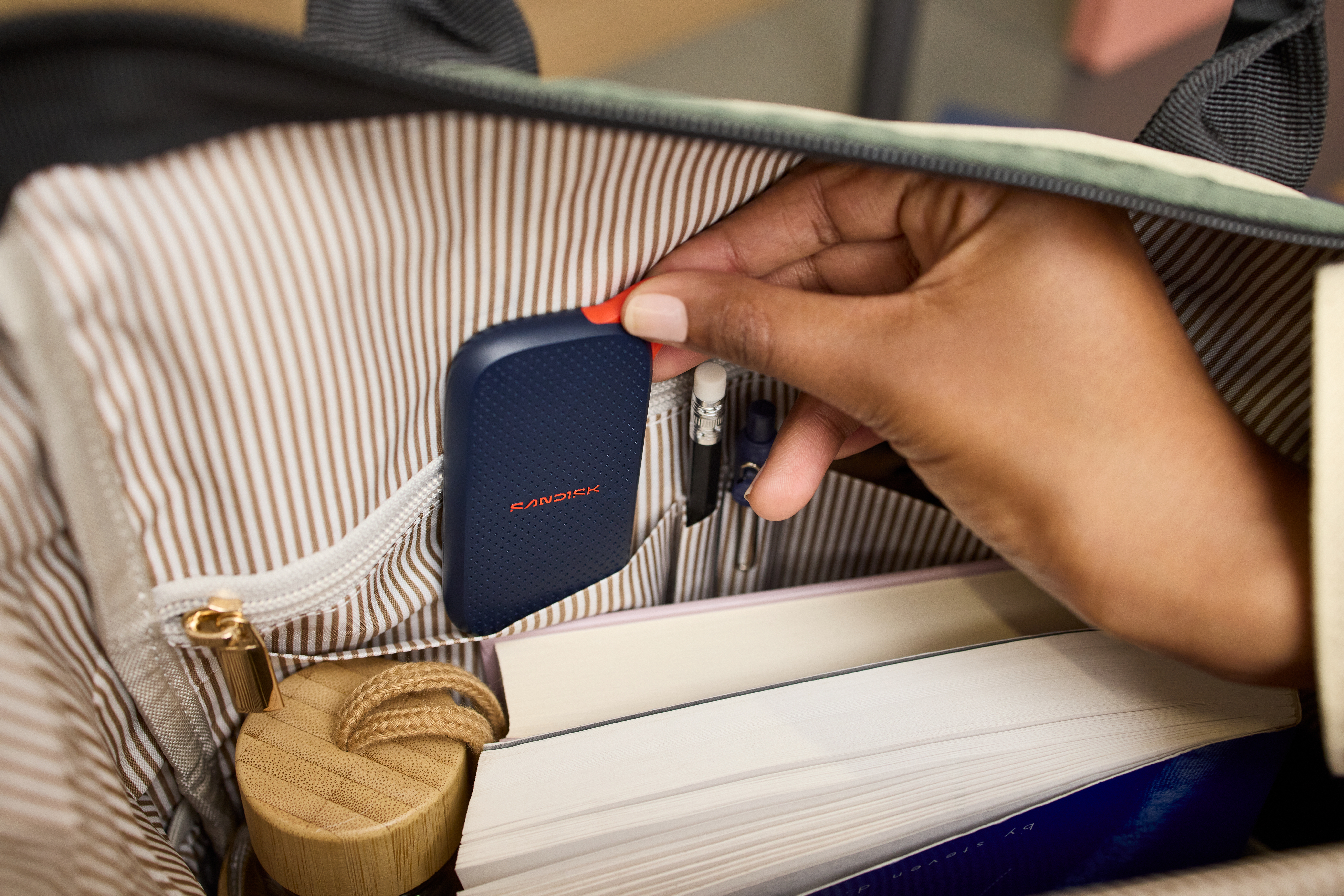 Hand legt dunkles blaues SanDisk-Laufwerk in eine Tasche mit Büchern und Holzgegenstand.