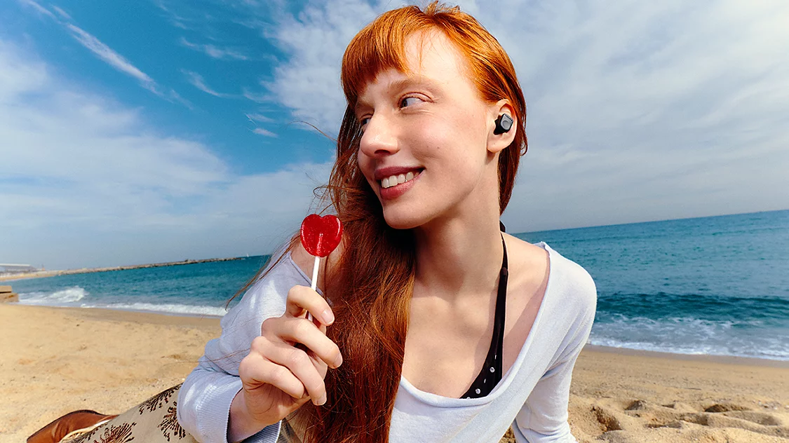 Eine Frau mit roten Haaren hält einen roten Herz-Lutscher am Strand.