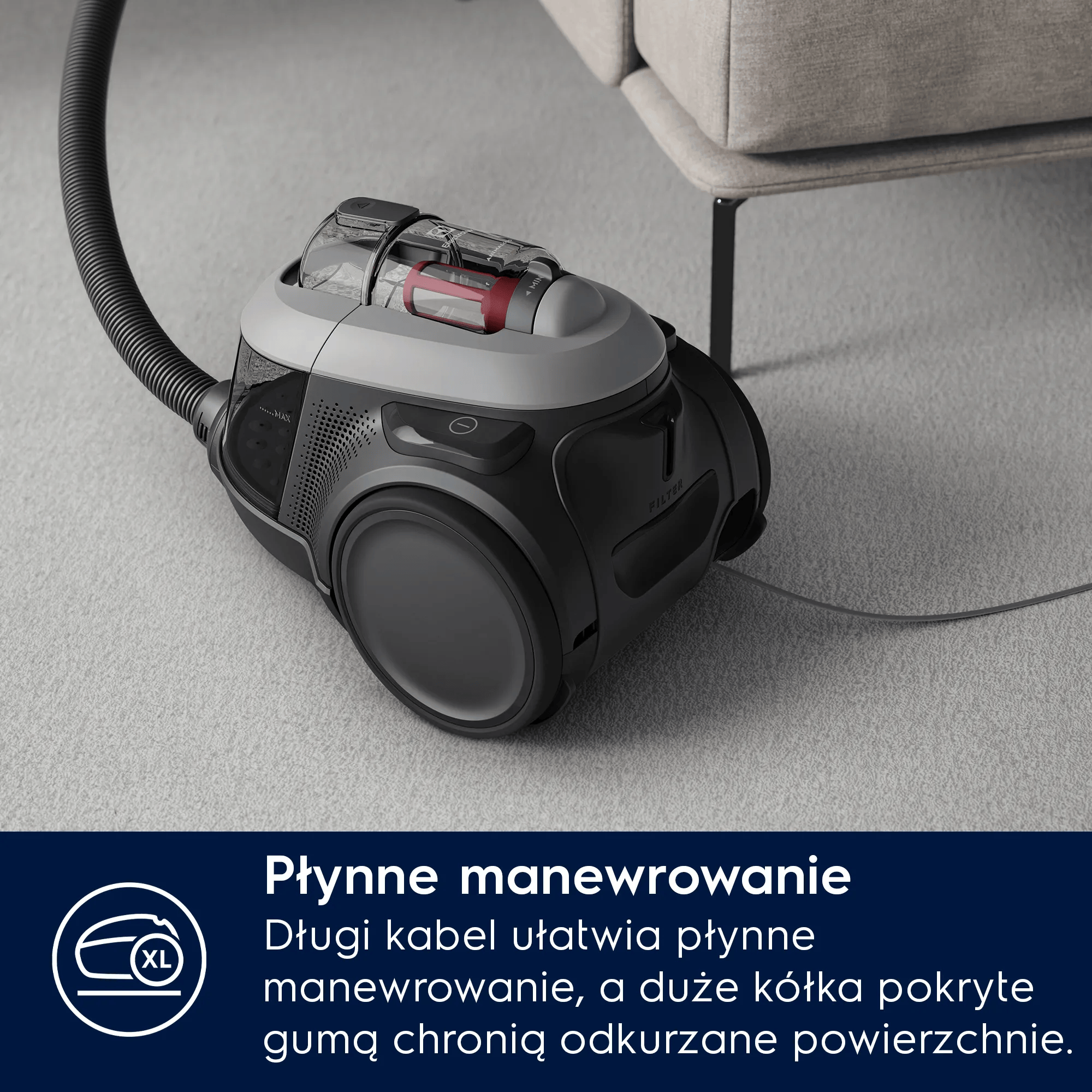 Szary odkurzacz z wężem stoi na dywanie obok kanapy.
