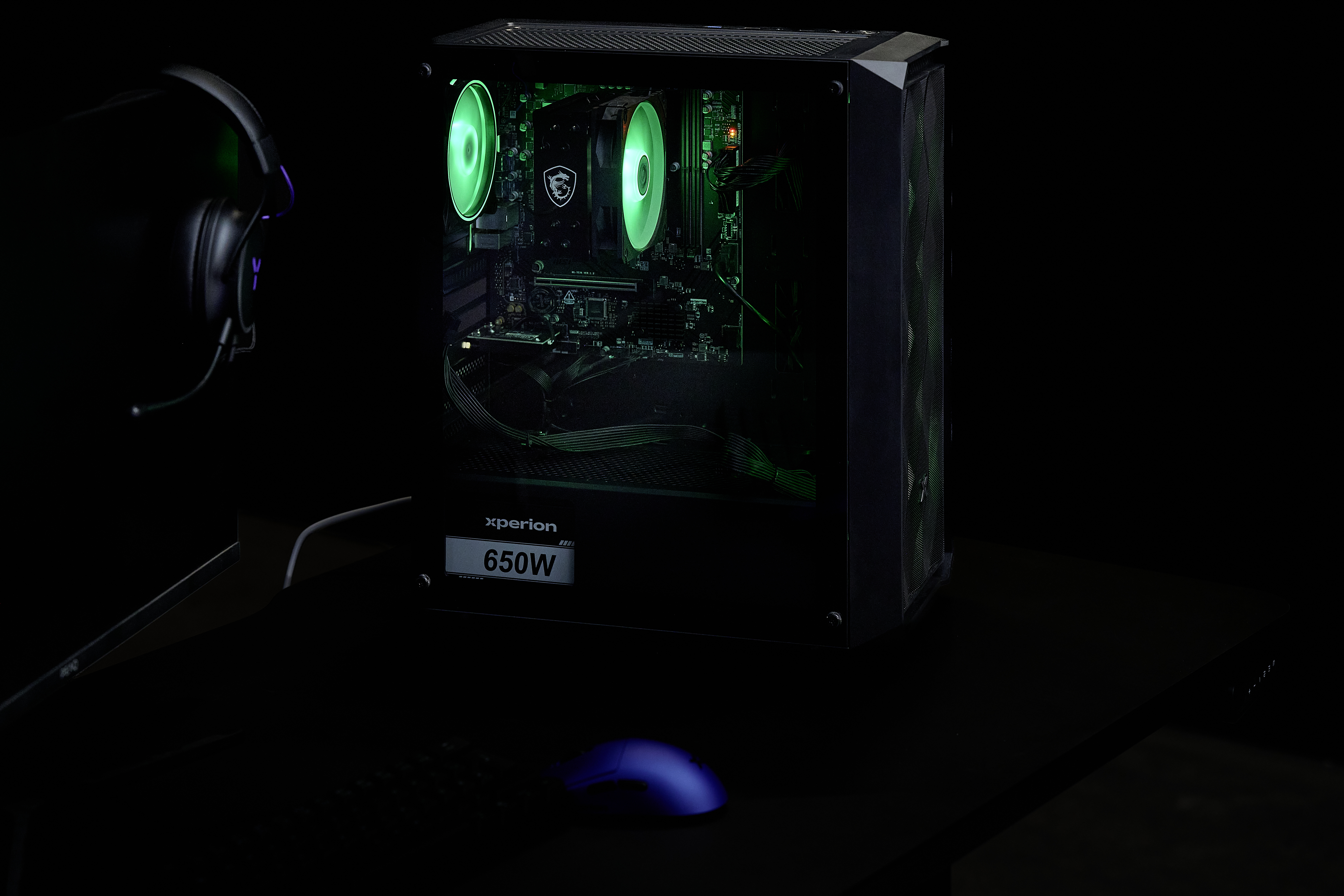 Ein schwarzer Gaming-PC mit grüner Beleuchtung, Kopfhörern und Maus auf einem Tisch.