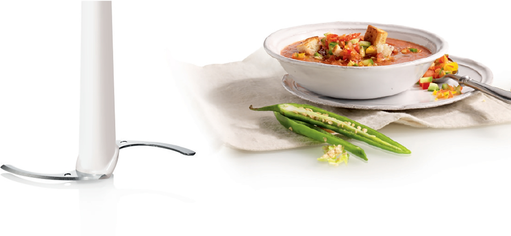 Ein Stabmixer mit Klingen neben einer Schüssel Gazpacho und zwei grünen Chilis.