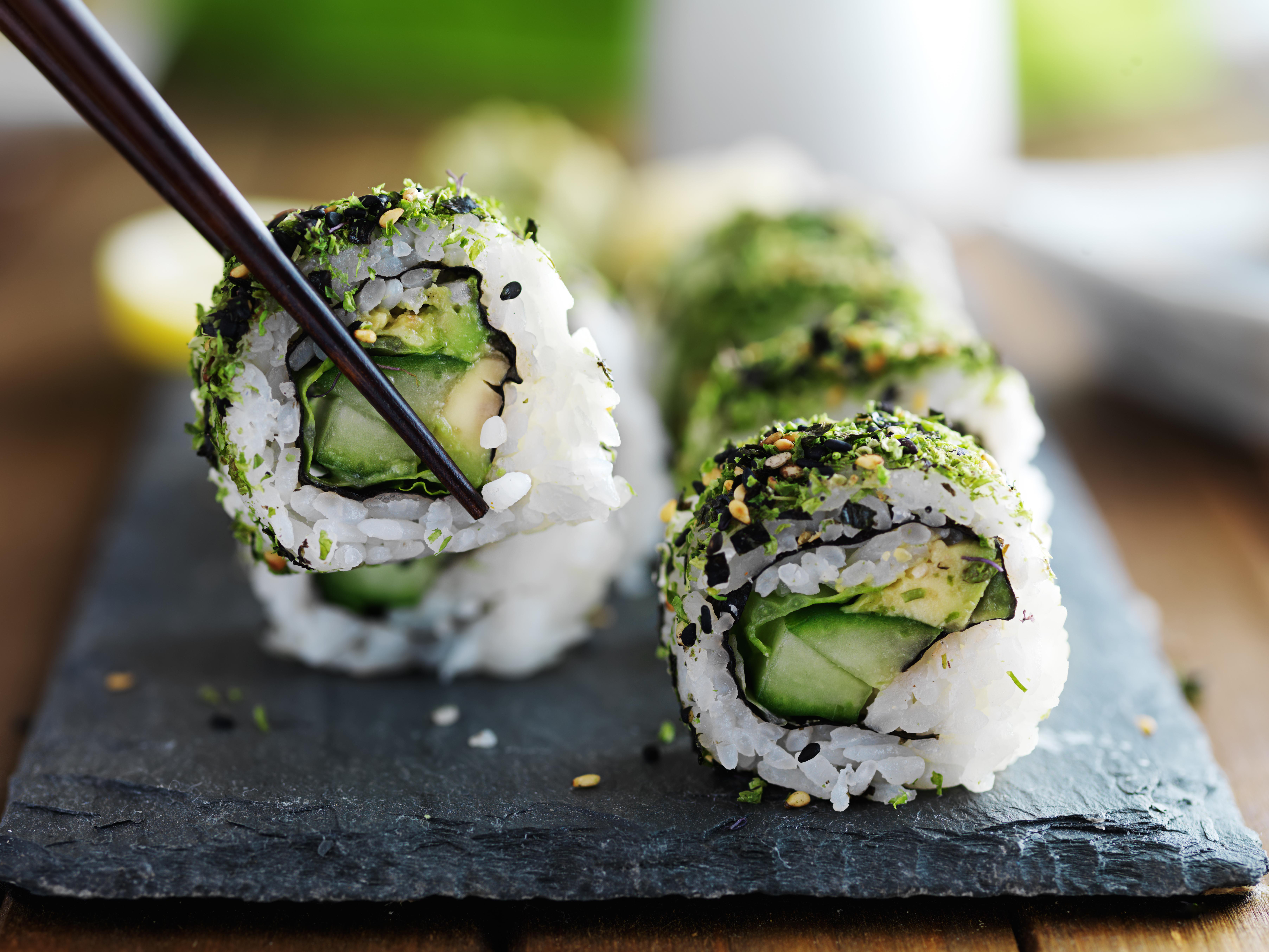 Sushi-Rolle mit Avocado und Gurke, bestreut mit Algen und Samen, wird mit Stäbchen gehalten.