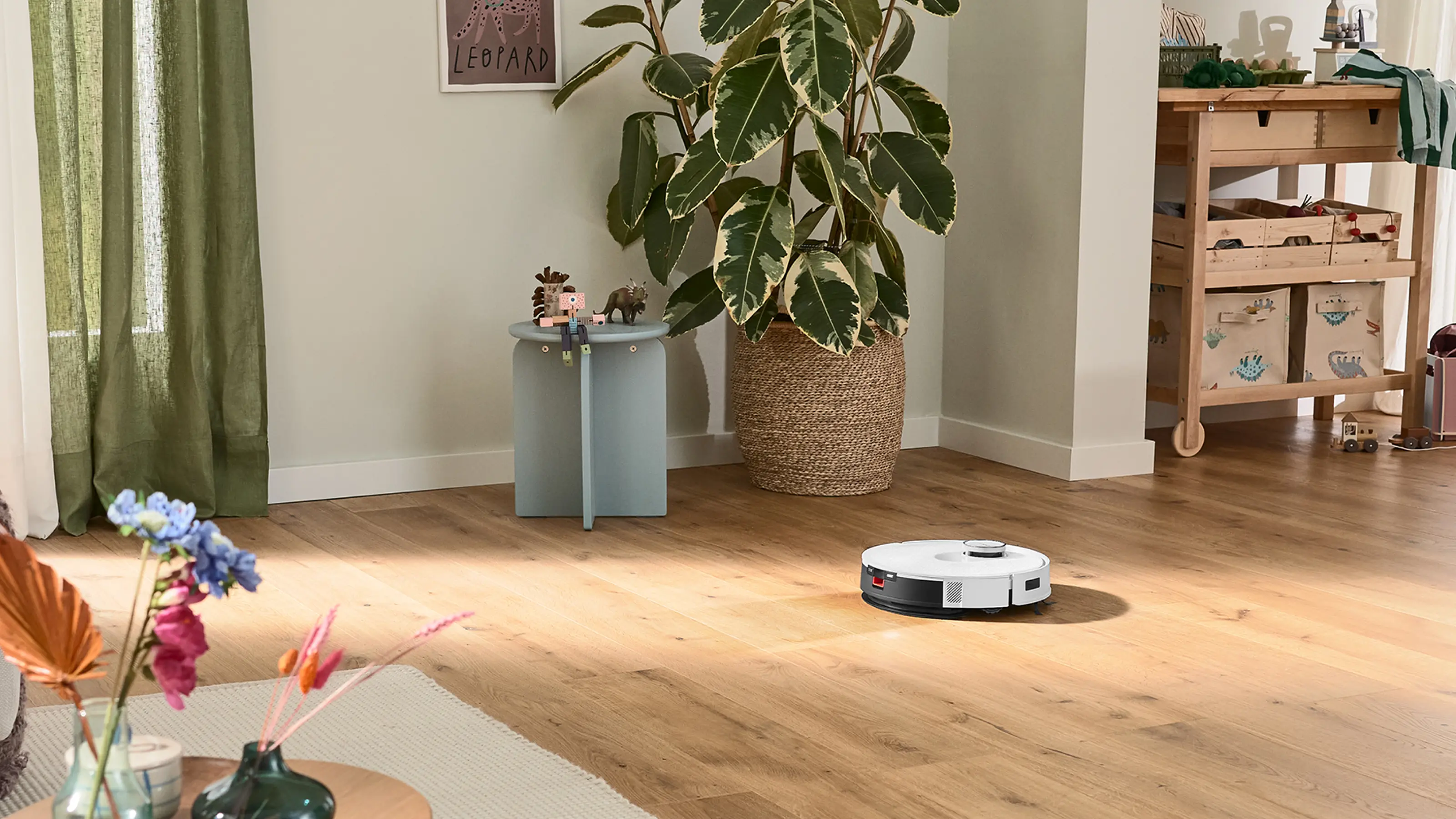 Egy fehér robotporszívó tisztít egy fapadlót. Növény és dekoráció látható.