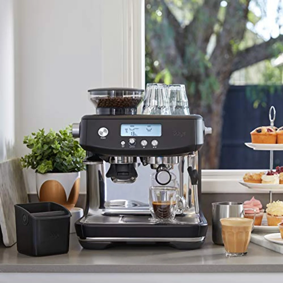 Una máquina de espresso negra con granos de café, vasos y tazas en una encimera.