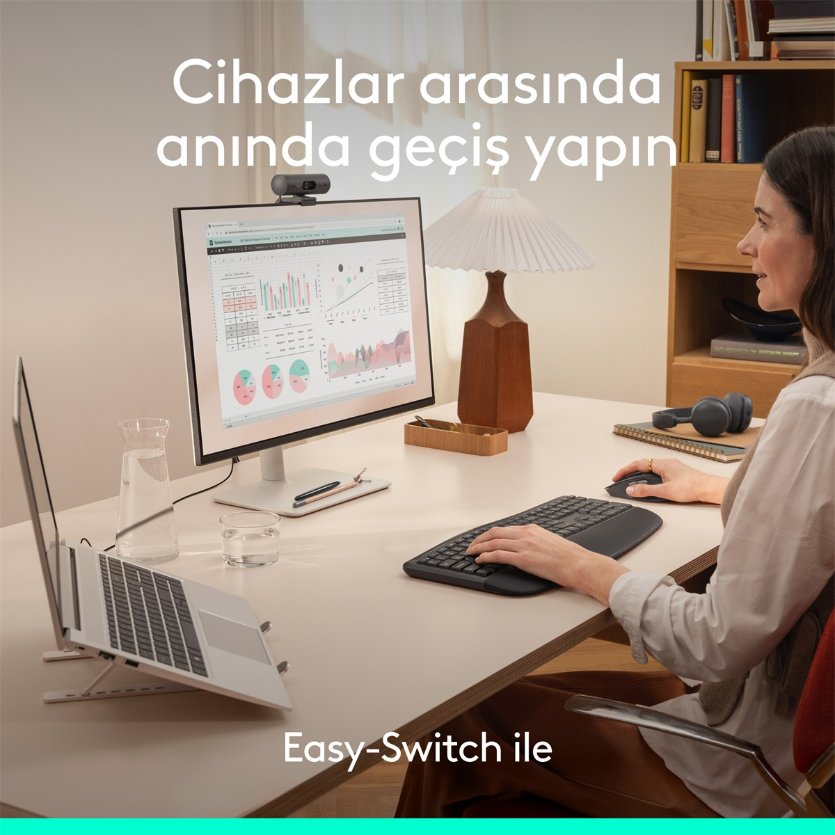 Bir kadın dizüstü bilgisayar, monitör ve klavyeyle masada çalışıyor. Kulaklıklar yakında.