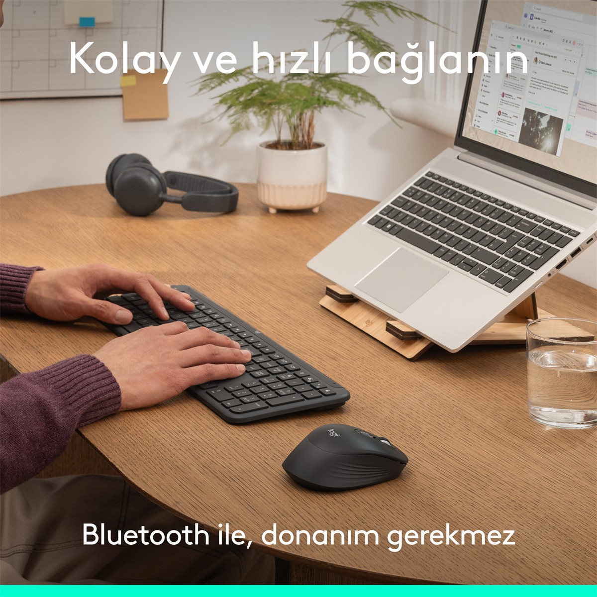 Kişi klavyede yazıyor, kulaklık, fare, stantta dizüstü bilgisayar, yanında su bardağı.