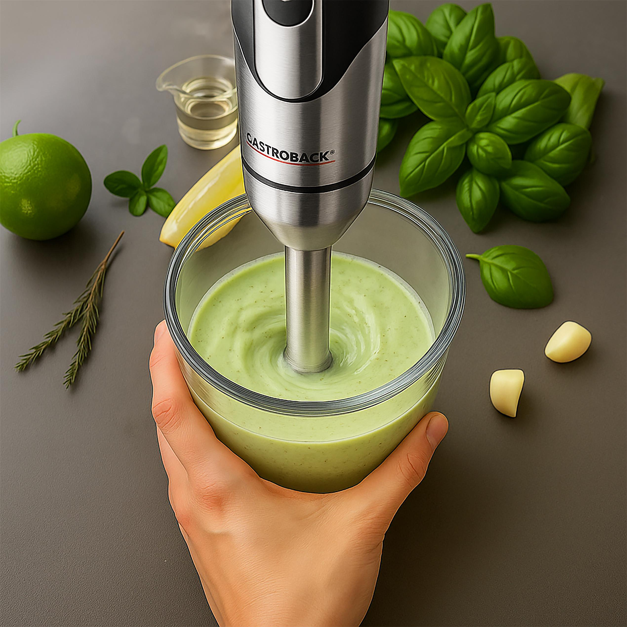 Ein Stabmixer mixt eine grüne Soße in einer Schüssel. Zutaten wie Basilikum, Limette und Knoblauch liegen daneben.
