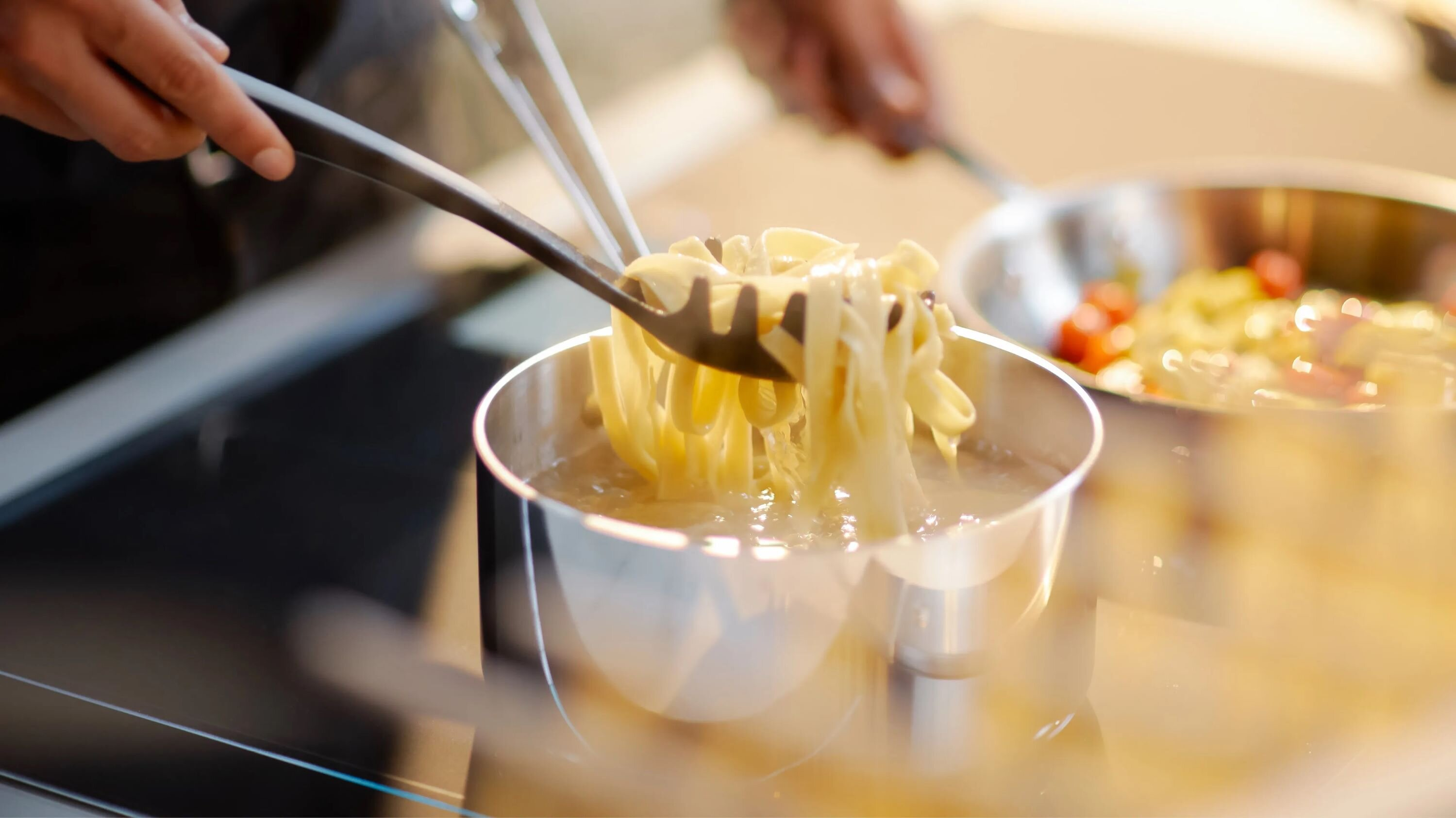 Makaron fettuccine unoszony z gotującej się wody szczypcami.
