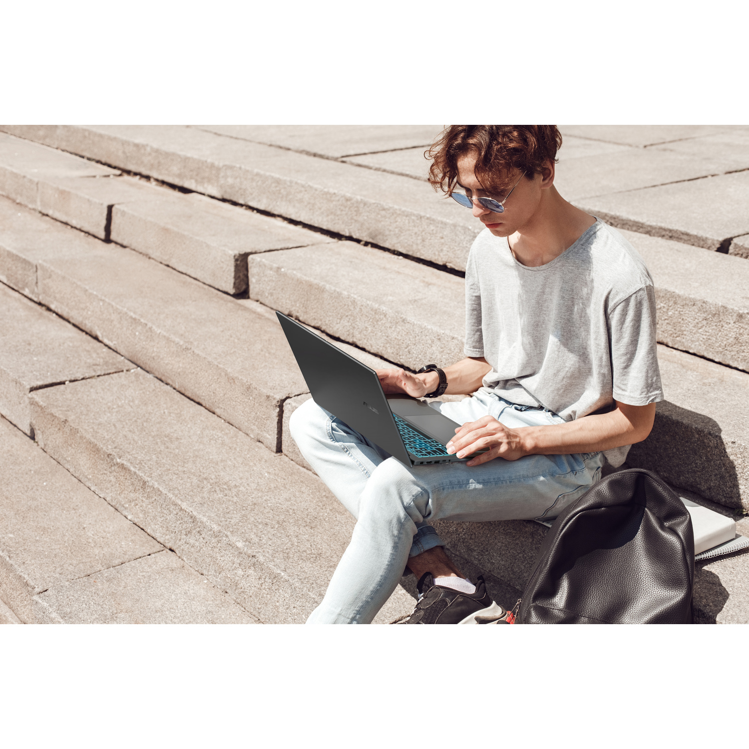 Un uomo seduto sui gradini di pietra usa un laptop. Indossa jeans e una maglietta.