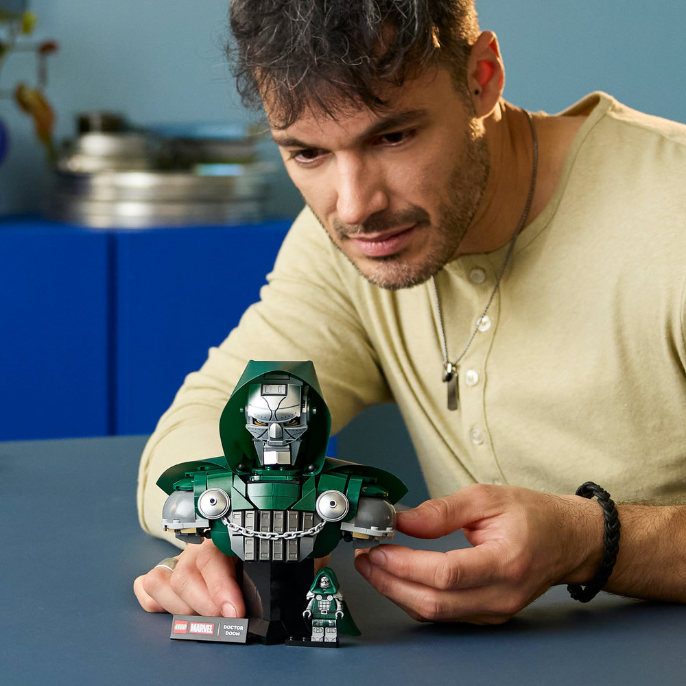 Ein Mann mit einem hellbeigen Hemd baut eine grüne Lego-Figur von Doctor Doom mit silbernen Details.
