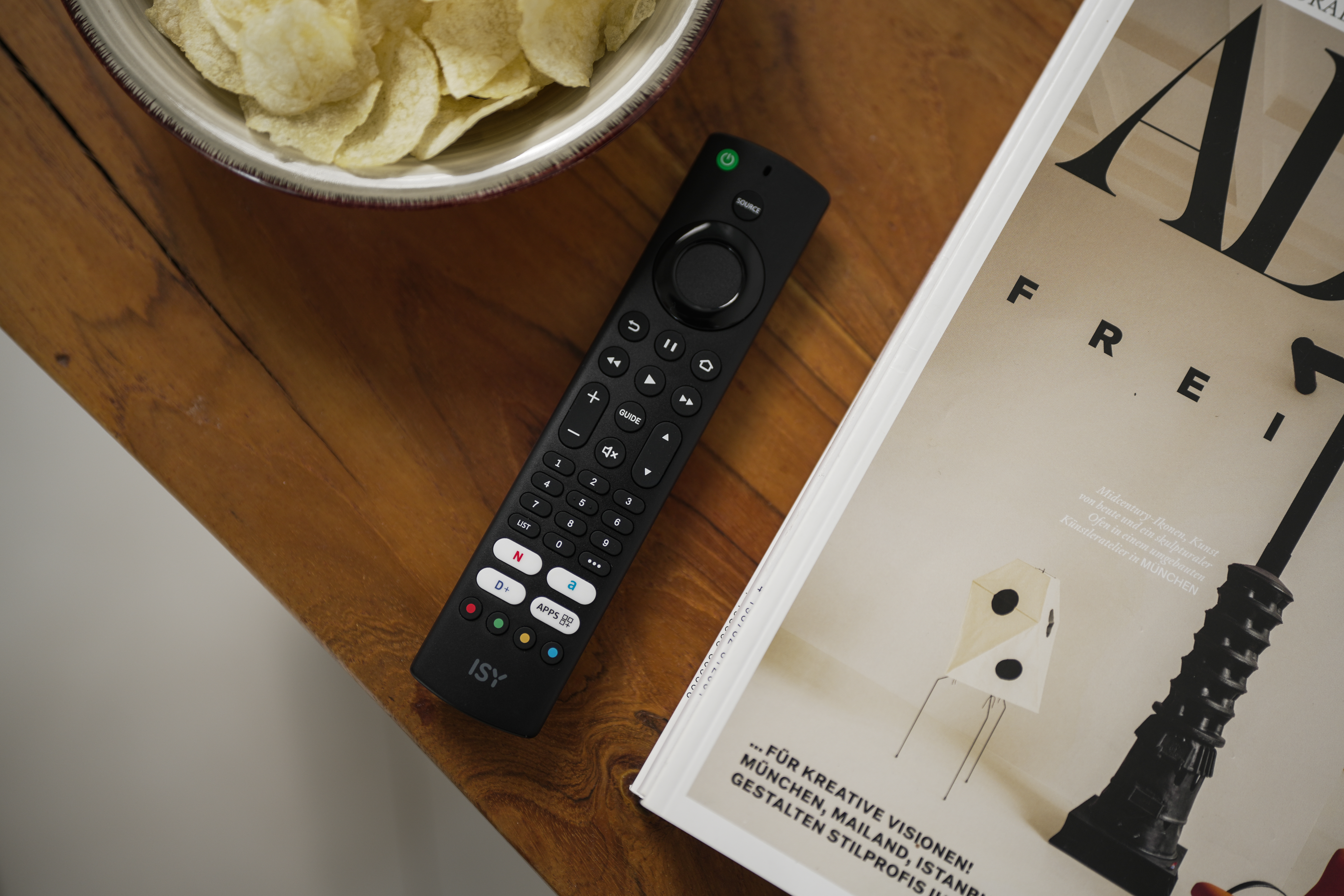 Schwarzer Fernbedienung neben Schüssel mit Chips und Magazin auf Holztisch.