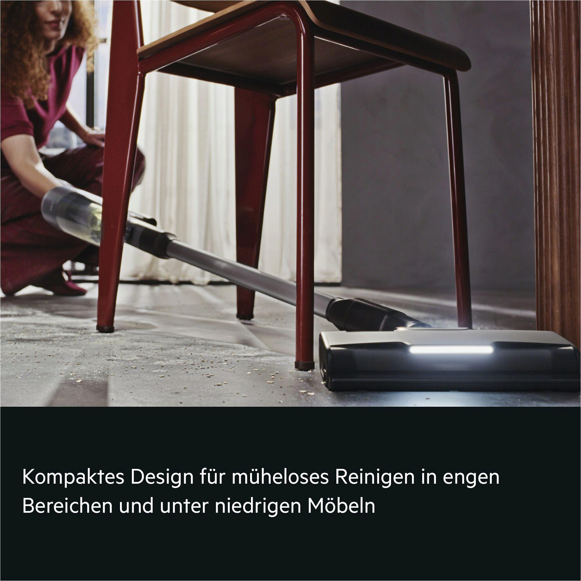 Eine Person saugt Staub unter einem roten Stuhl mit einem Handstaubsauger.