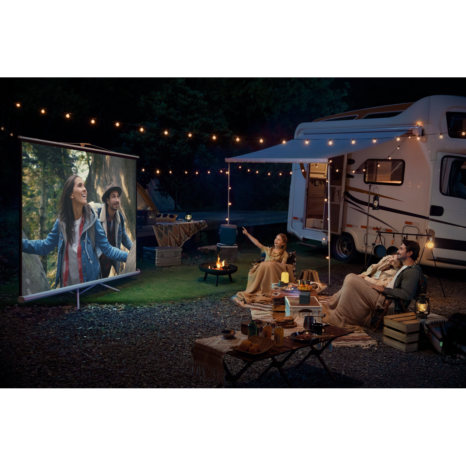 Couple campant la nuit devant un camping-car, regardant un film sur écran.