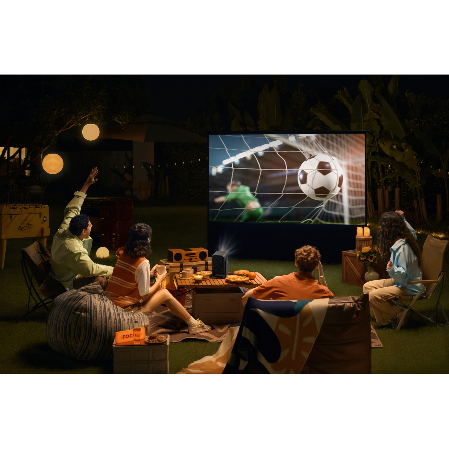 Des gens regardent du football en plein air avec un projecteur. Plusieurs personnes assises sur des couvertures et des coussins.