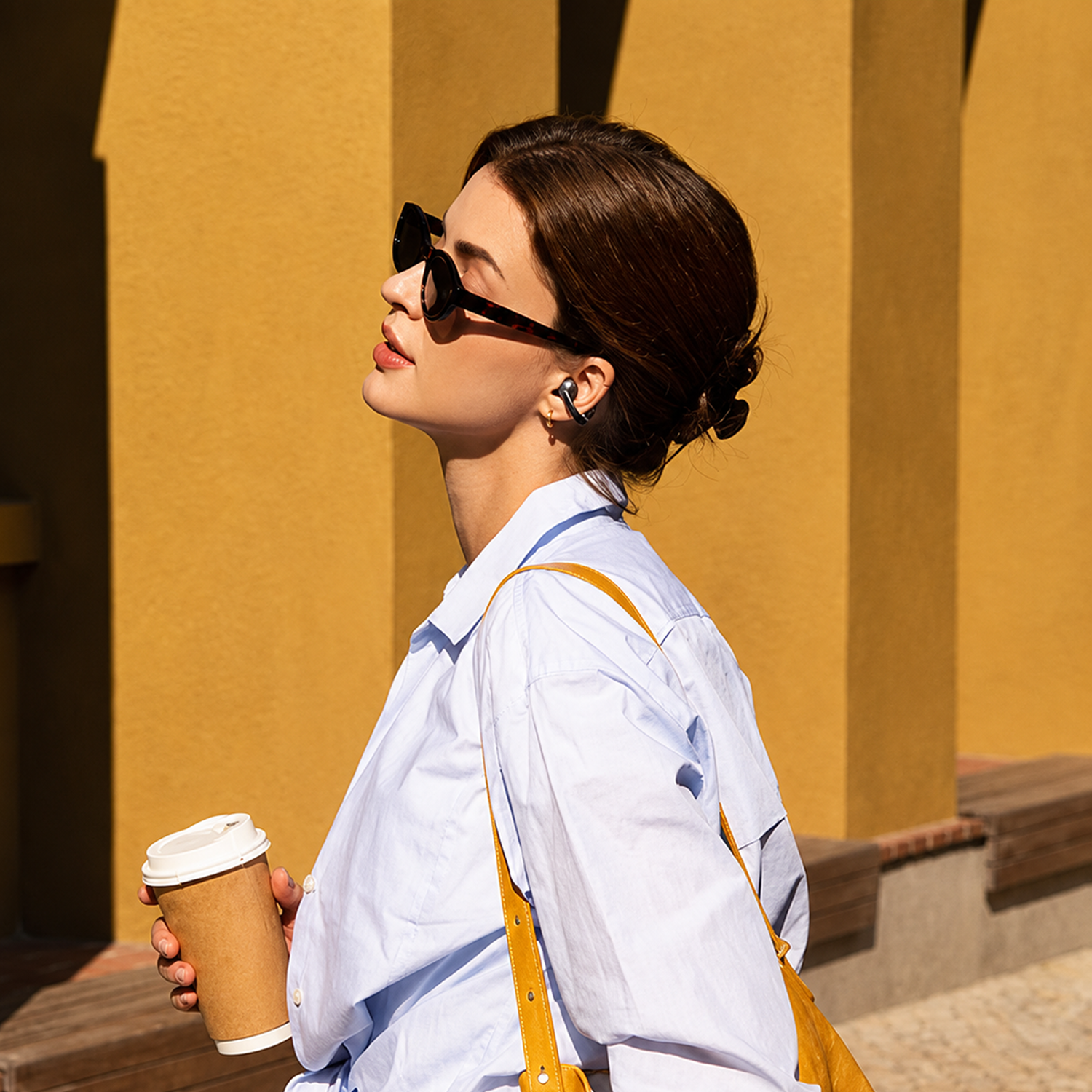Mujer con gafas de sol y audífonos sostiene taza de café, pared amarilla.