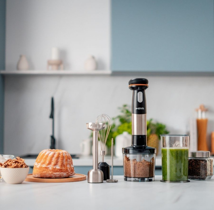 Blender ręczny z akcesoriami obok ciasta, orzechów i zielonego smoothie.