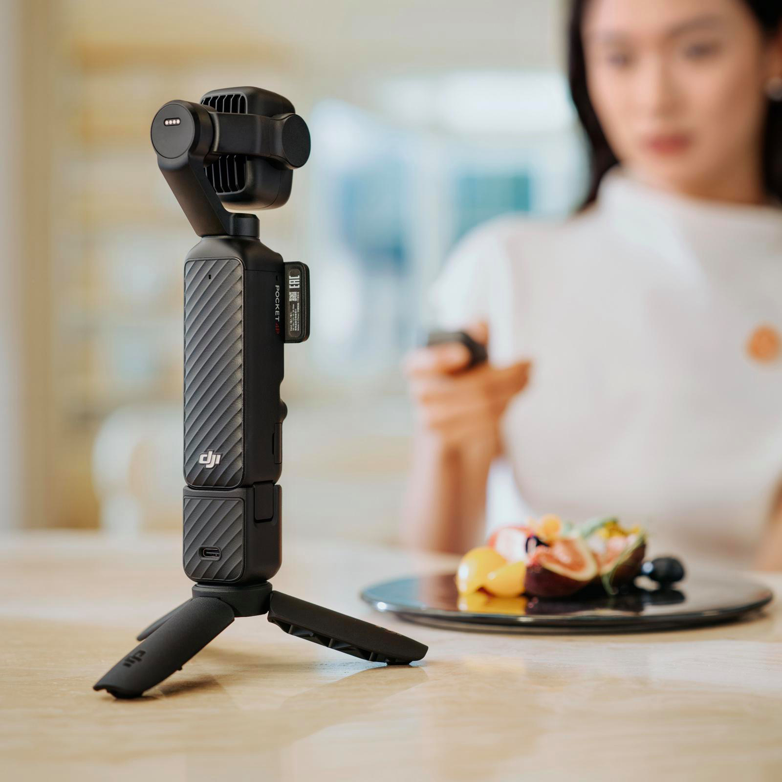 Ein schwarzer Gimbal mit Stativ steht auf einem Tisch. Eine Frau ist im Hintergrund unscharf.