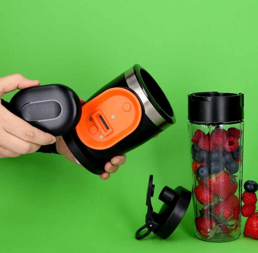 Ręka trzyma czarny kubek do smoothie z pomarańczową podstawą. Obok szklany kubek z jagodami.
