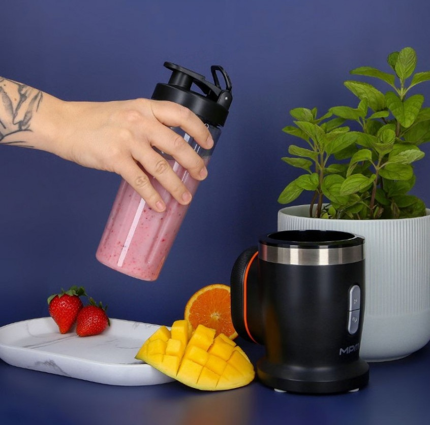 Ręka trzyma butelkę blendera z smoothie obok owoców i blendera.