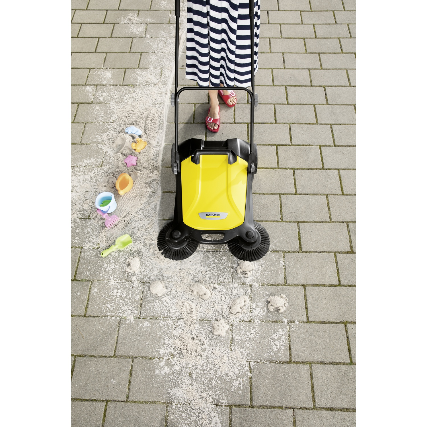 Una spazzatrice Karcher gialla viene spinta da una persona con vestito a righe e sandali rossi su un patio.