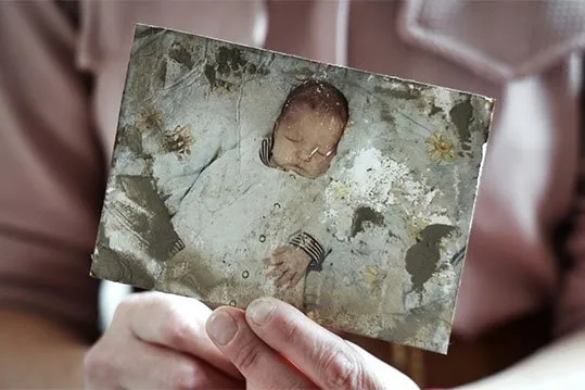 Una foto danneggiata di un bambino addormentato in una tutina bianca, tenuta dalle mani.