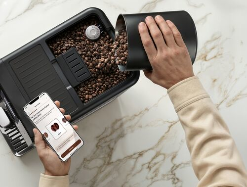 Une main verse des grains de café dans une machine à café tandis qu'une autre tient un smartphone.
