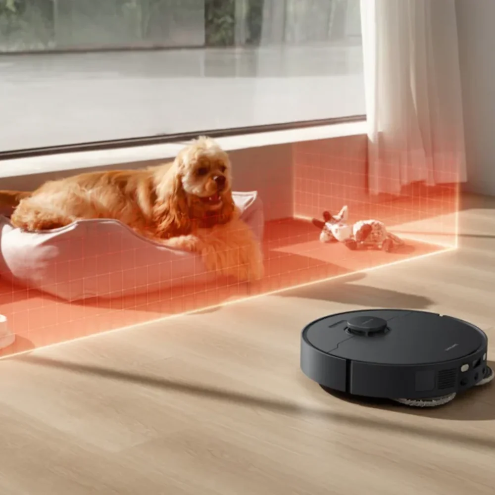 Egy robotporszívó tisztítja a padlót egy kutya mellett, amely az ágyában pihen.