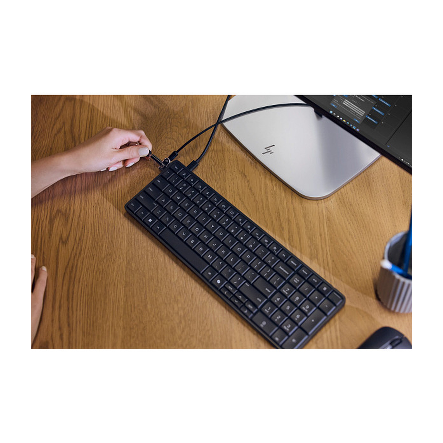 Una mano inserisce un cavo USB nero in una tastiera nera. Si vedono monitor e mouse.