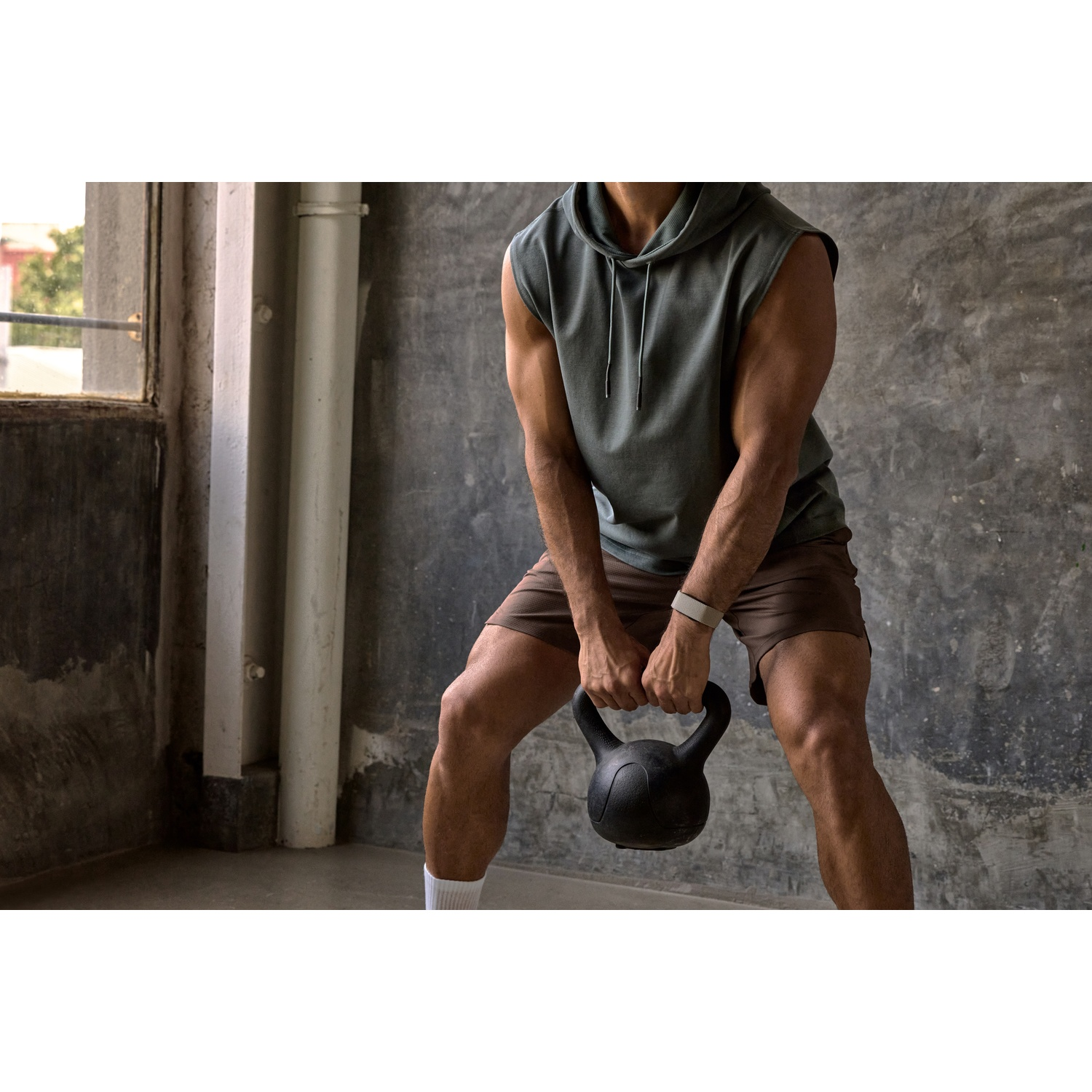 Man tilt kettlebell in de sportschool, draagt grijze mouwloze hoodie en bruine korte broek.