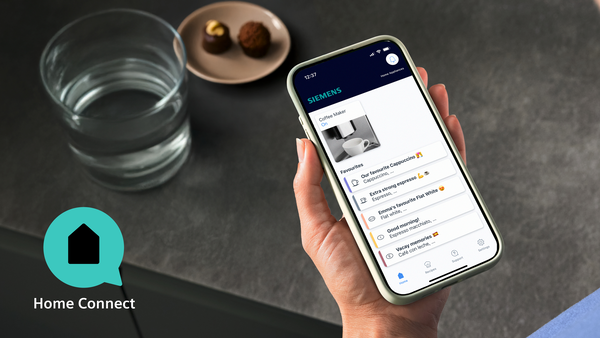 Bir el Home Connect uygulamalı akıllı telefon tutuyor. Yanında bir bardak su ve çikolatalar.