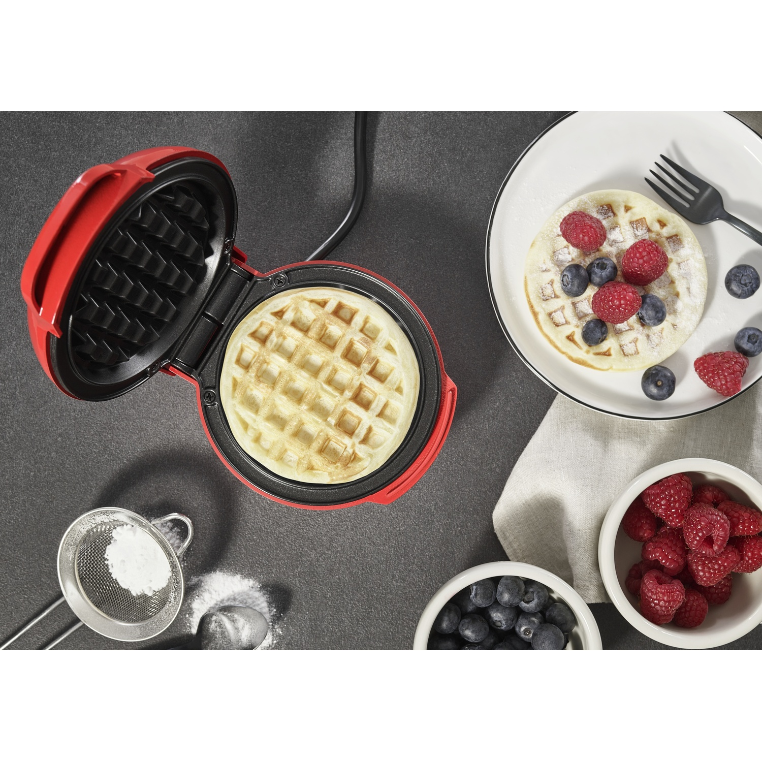 Una piastra per waffle rossa con un waffle all'interno, accanto a un piatto con un waffle, frutti di bosco e una forchetta.
