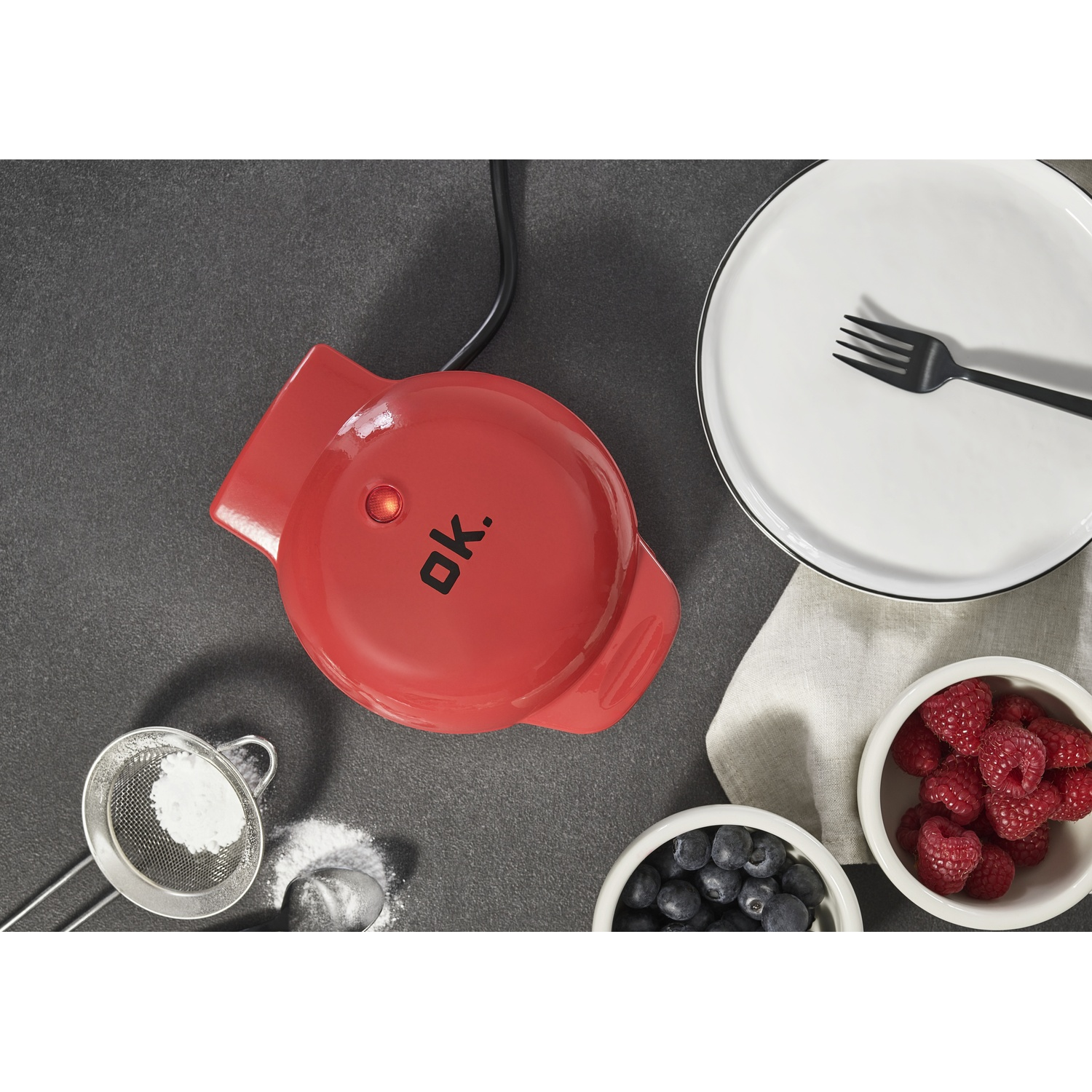 Waffle maker rosso con bacche, zucchero a velo e forchetta su piatto.