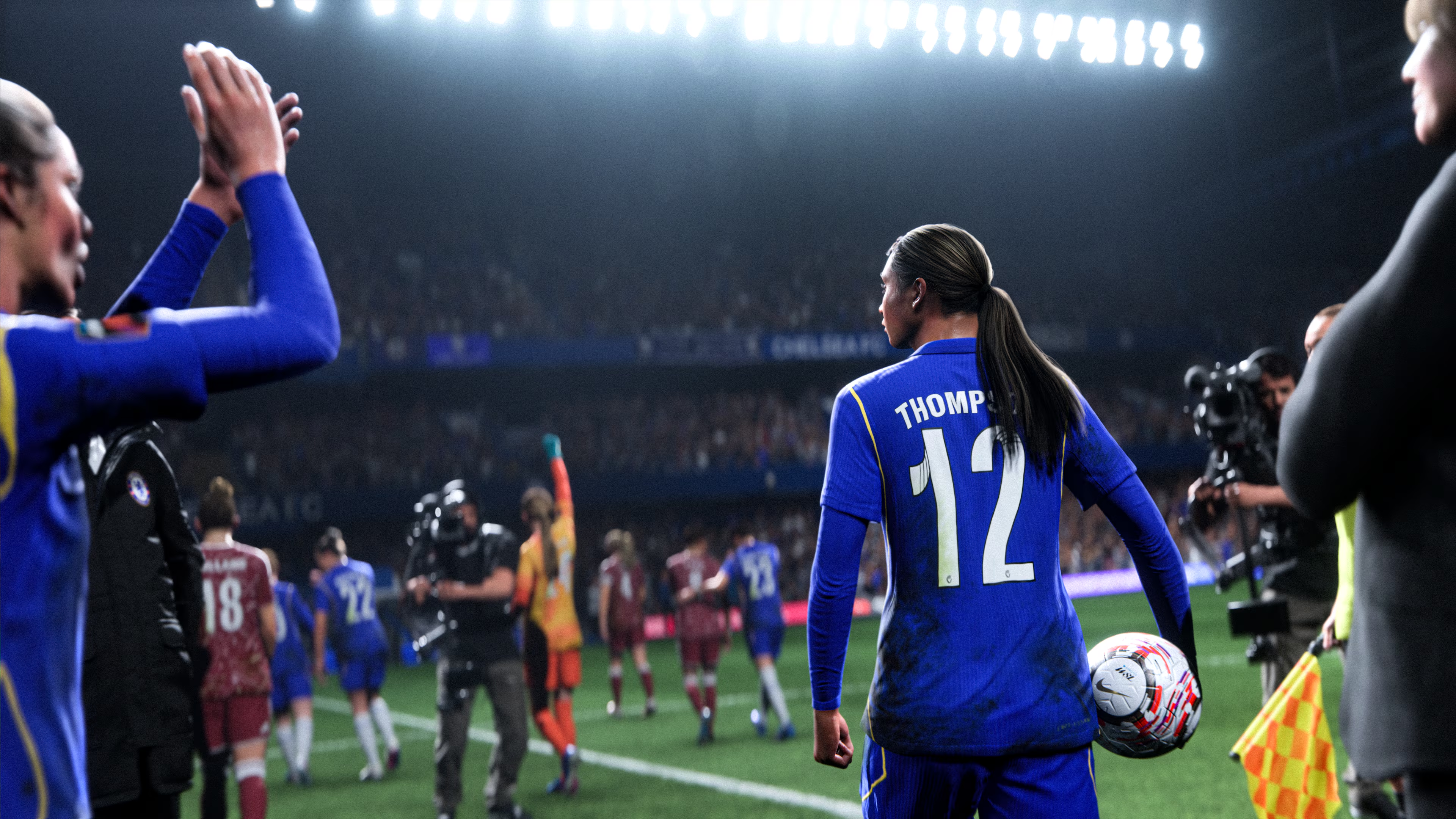 Calciatrice in maglia blu n. 12 tiene il pallone. Altre giocatrici sullo sfondo.