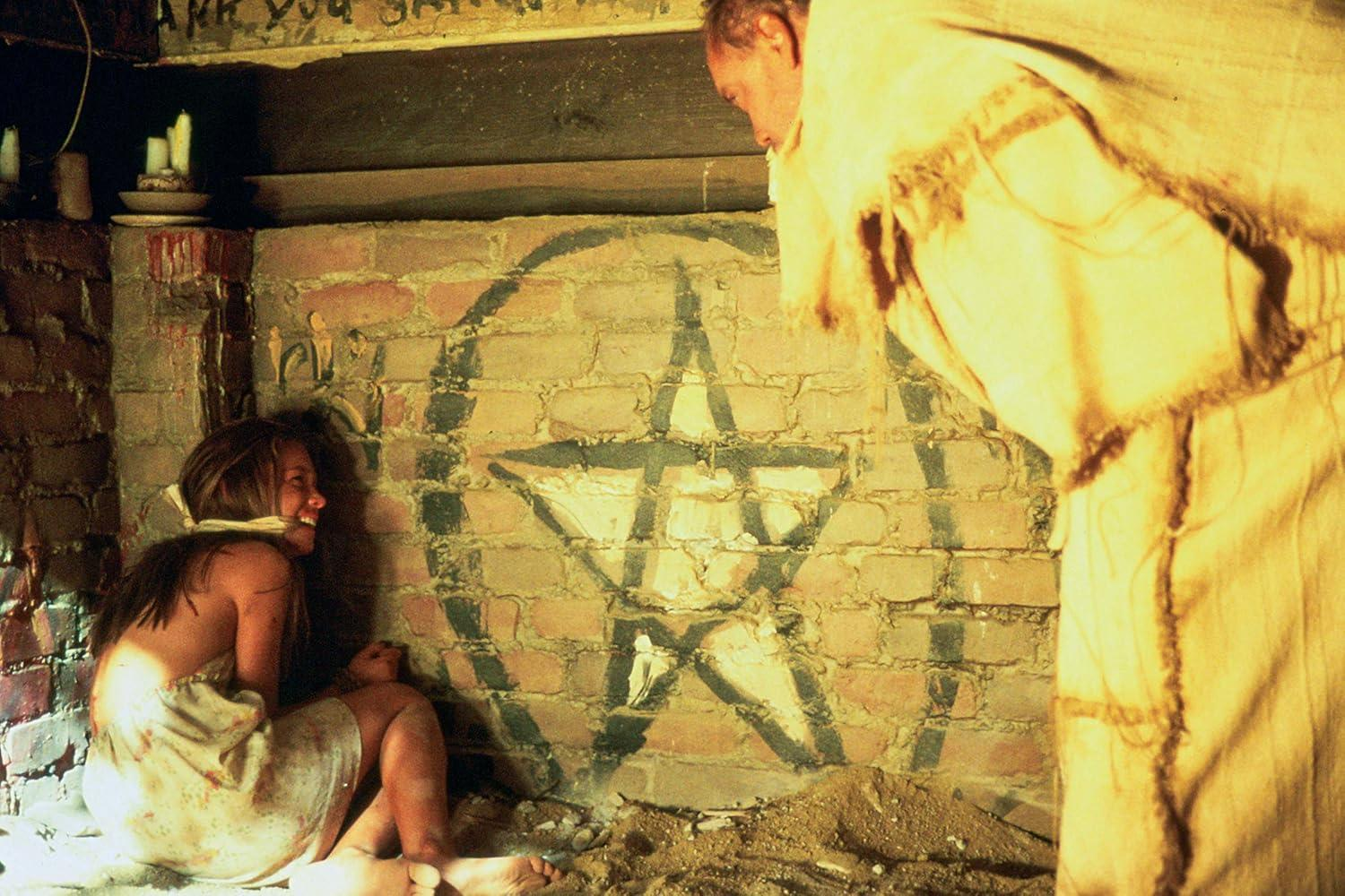 Eine gefesselte Frau sitzt auf dem Boden vor einer Wand mit einem aufgemalten Pentagramm, während ein Mann sie beobachtet.
