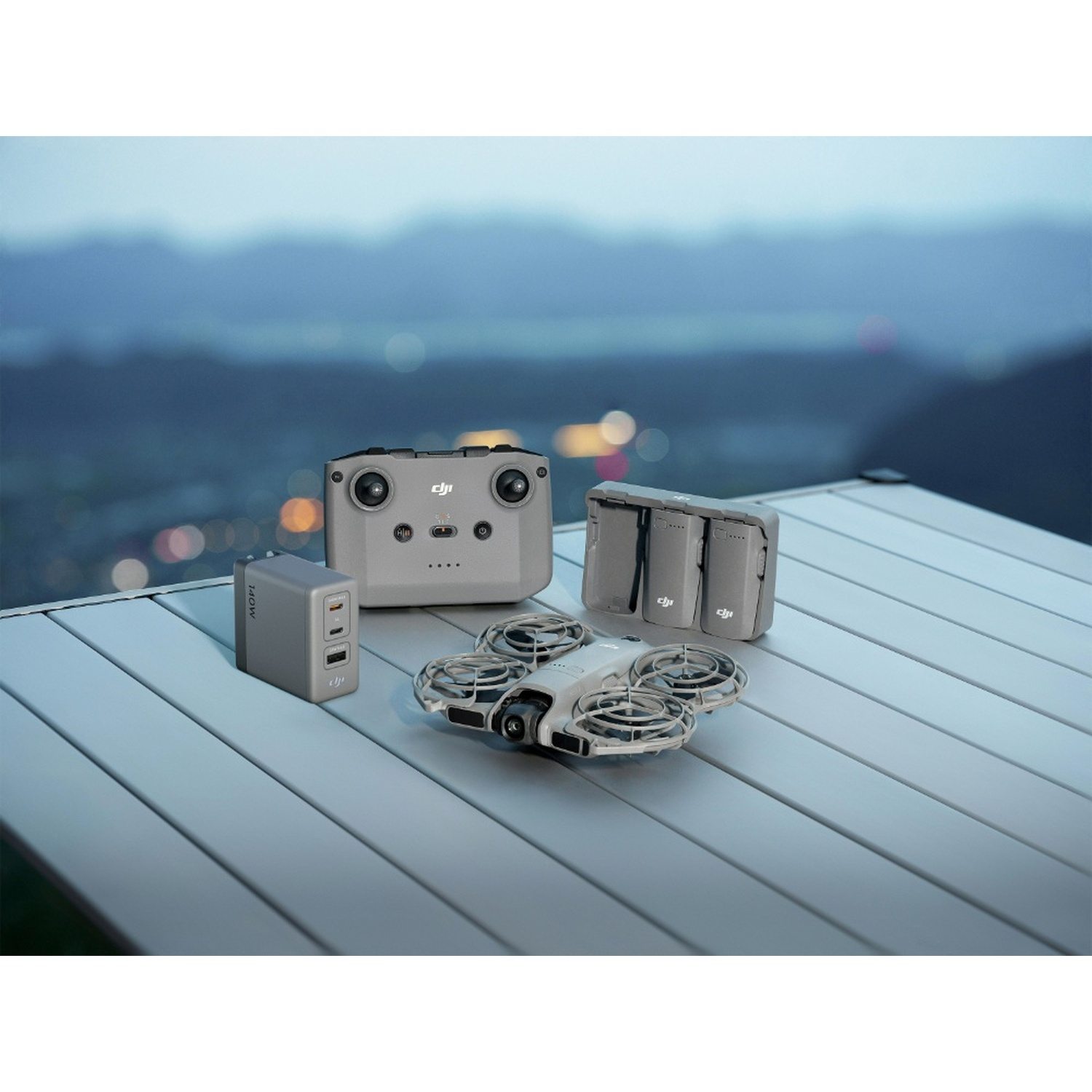 Un drone grigio con telecomando, batterie e caricabatterie su un tavolo con paesaggio montano sfocato sullo sfondo.