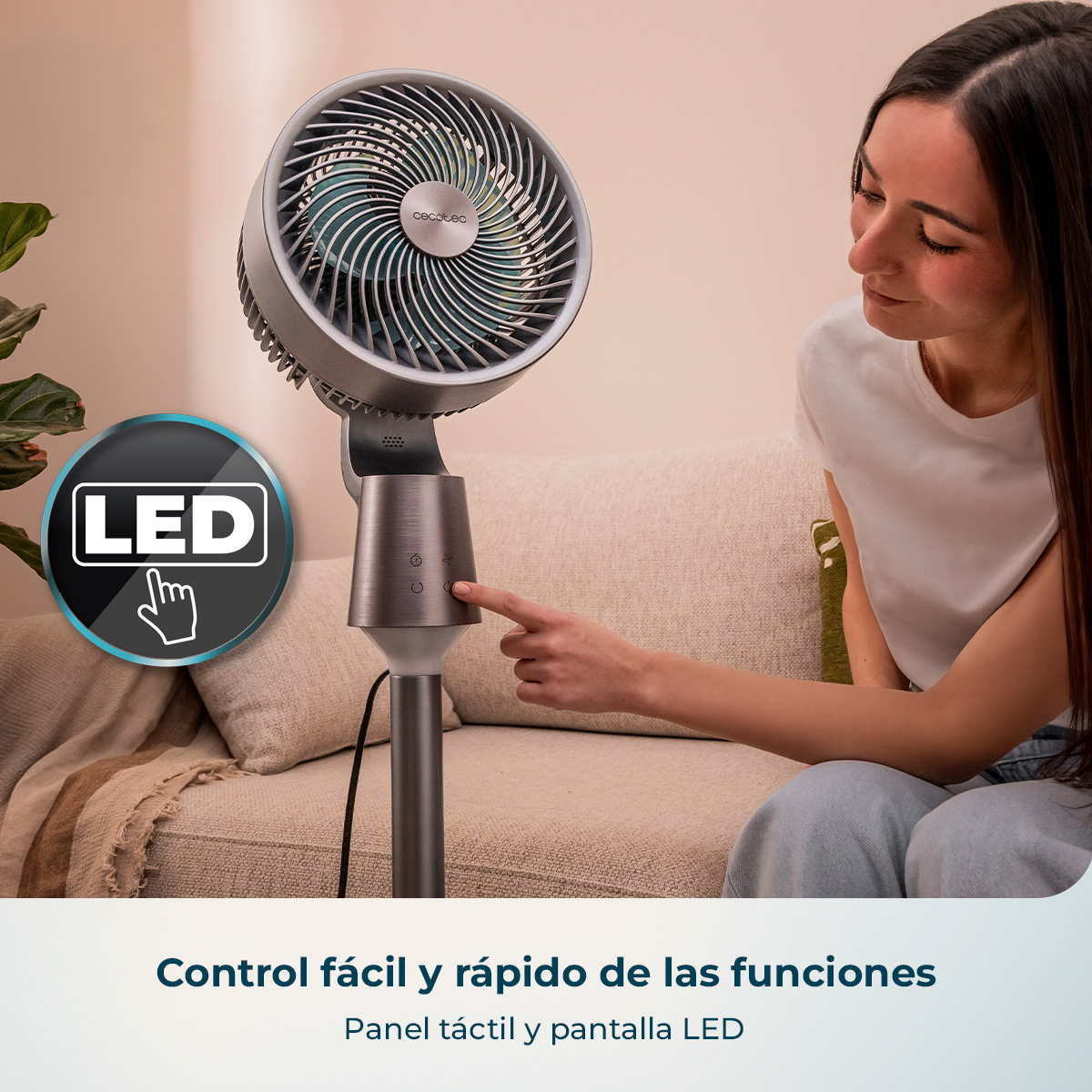 Una mujer está sentada en un sofá operando el panel táctil de un ventilador de pie con pantalla LED.