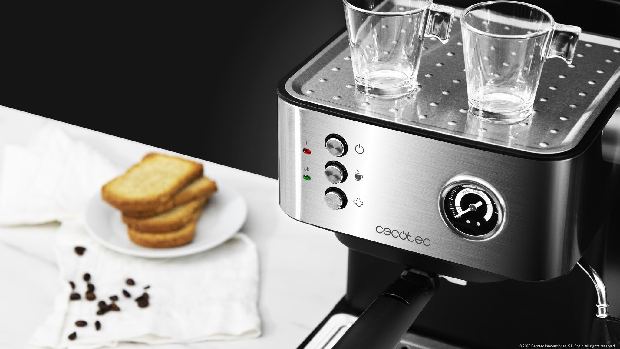 Una cafetera de acero inoxidable con dos tazas encima, botones, un manómetro y tostadas sobre un paño blanco.