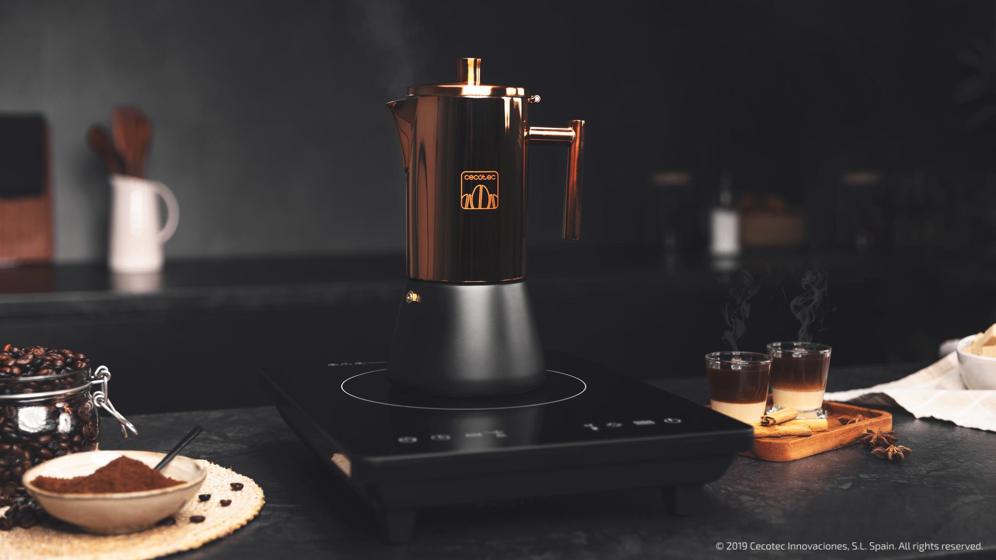 Cafetera italiana de cobre y negro sobre una placa de inducción, con café molido y vasitos al lado.