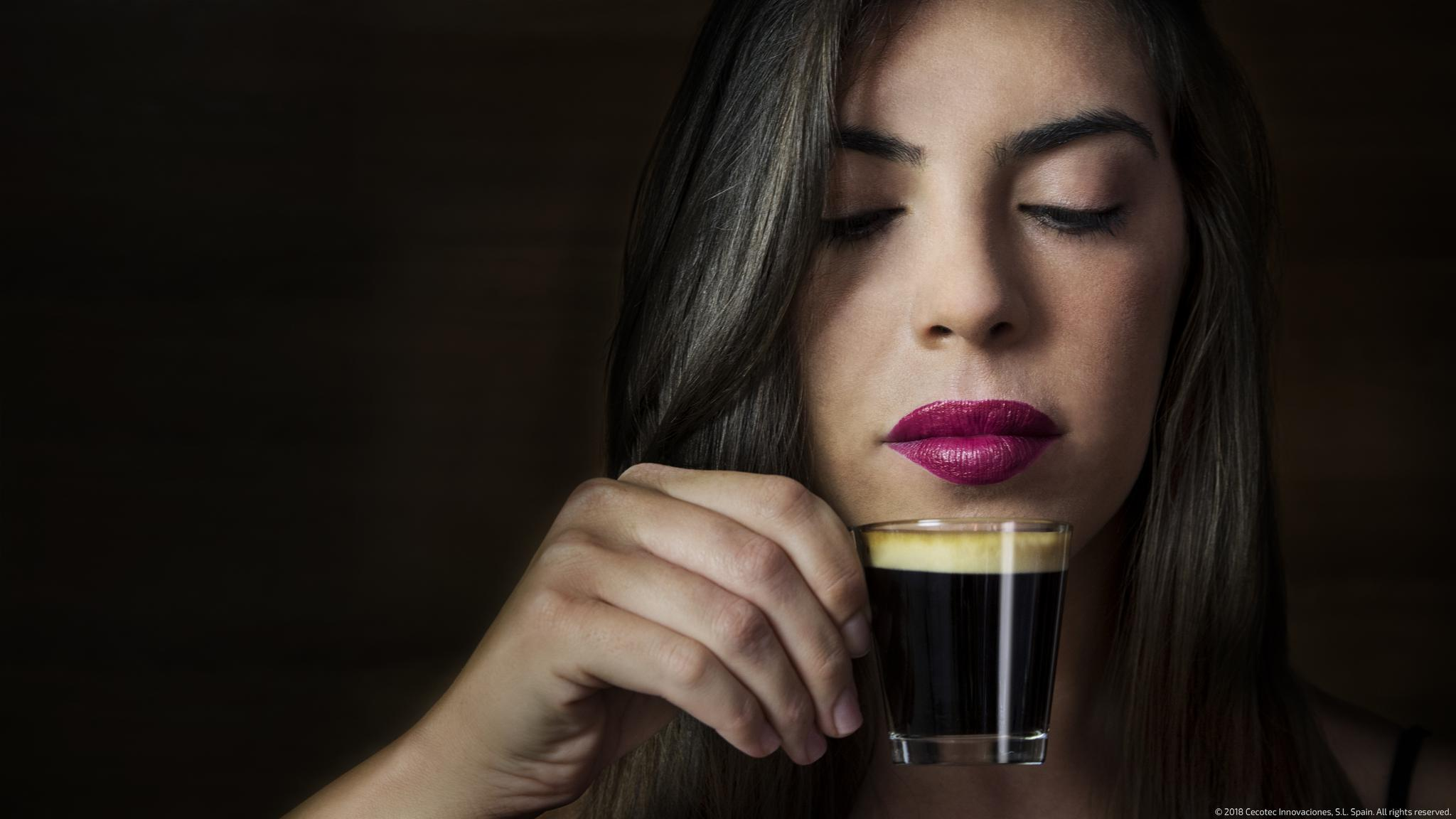 Una mujer con labios pintados de rosa sostiene una taza de espresso y la huele con los ojos cerrados.