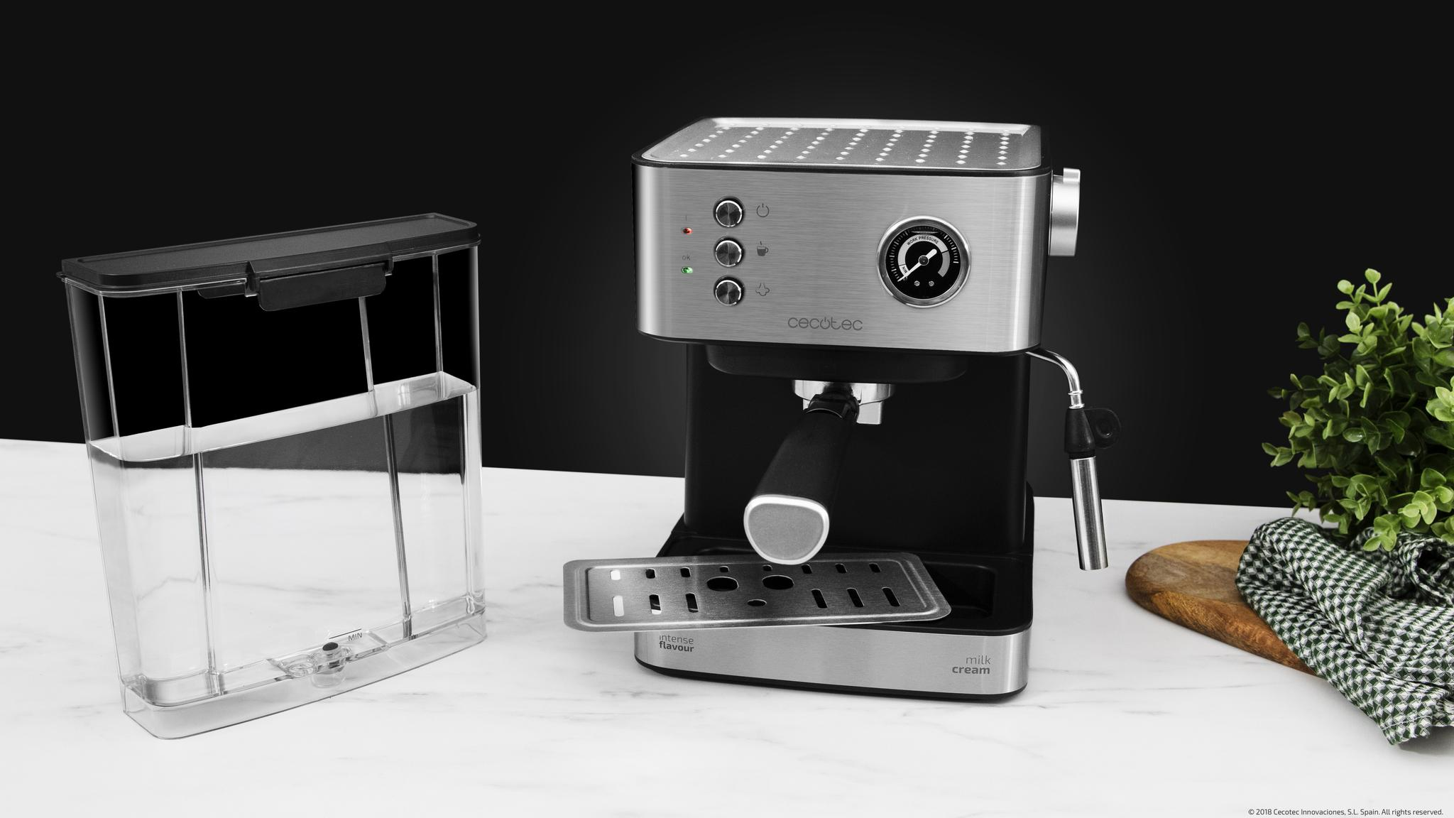 Una cafetera plateada con depósito de agua extraíble sobre una encimera blanca junto a una pequeña planta verde.