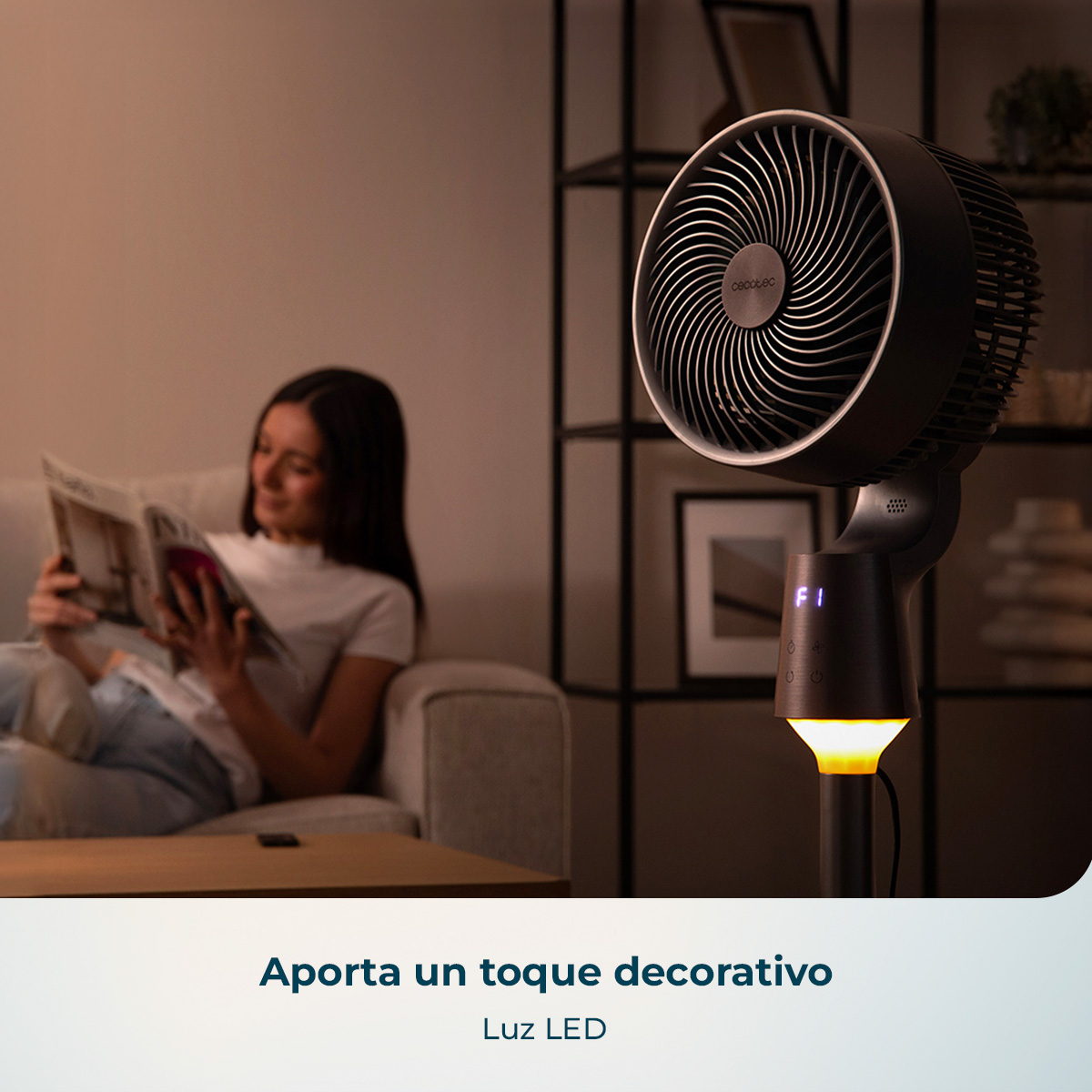 Un ventilador de pie negro con luz LED cálida en la base en una habitación mientras una mujer lee en un sofá.