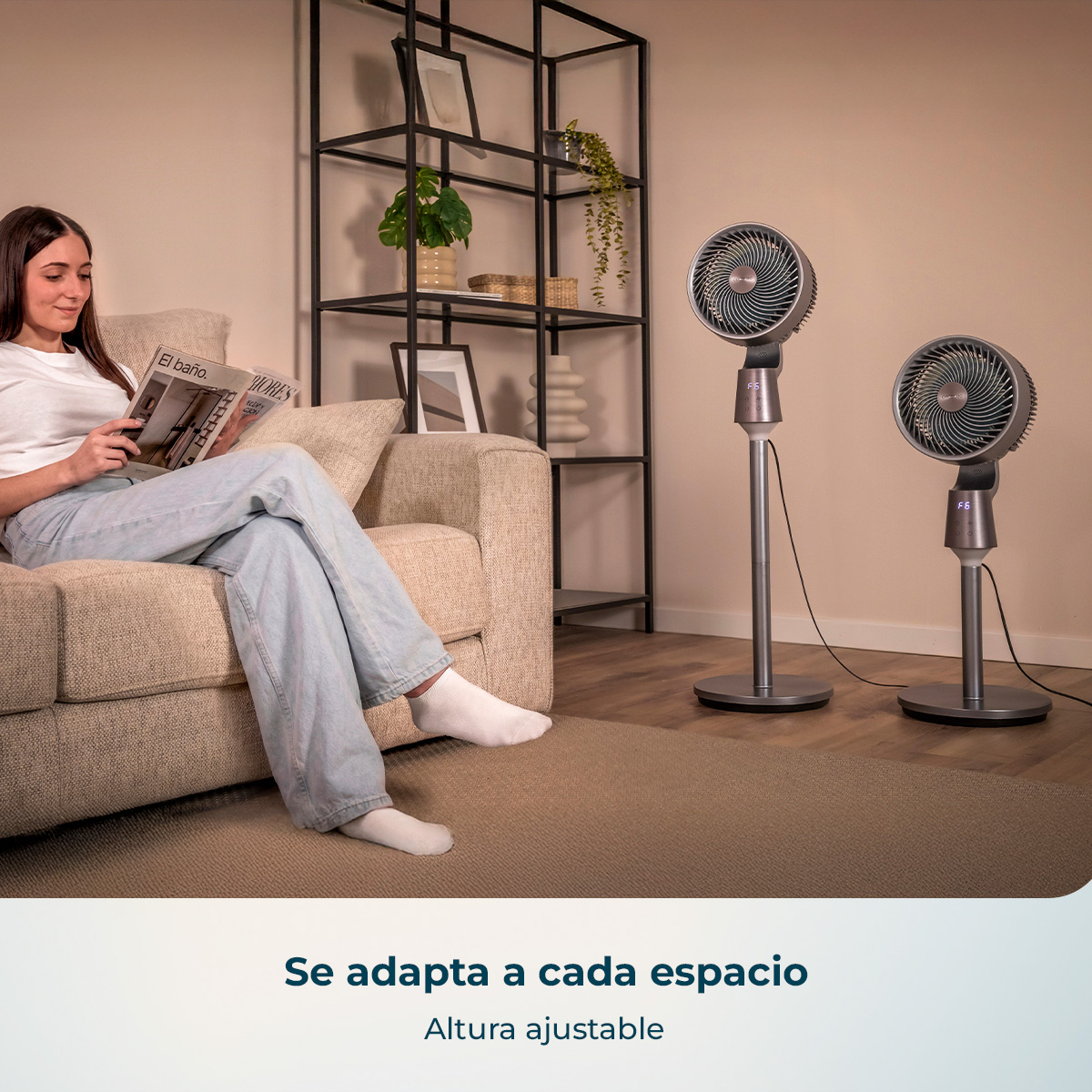 Una mujer lee en un sofá con dos ventiladores de pie grises en la habitación.