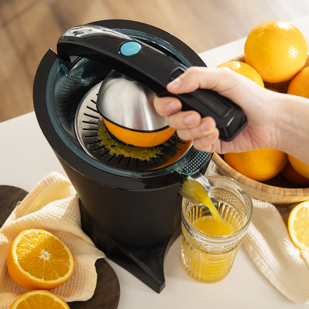 Una mano exprime una naranja en un exprimidor negro, el jugo fresco fluye a un vaso.