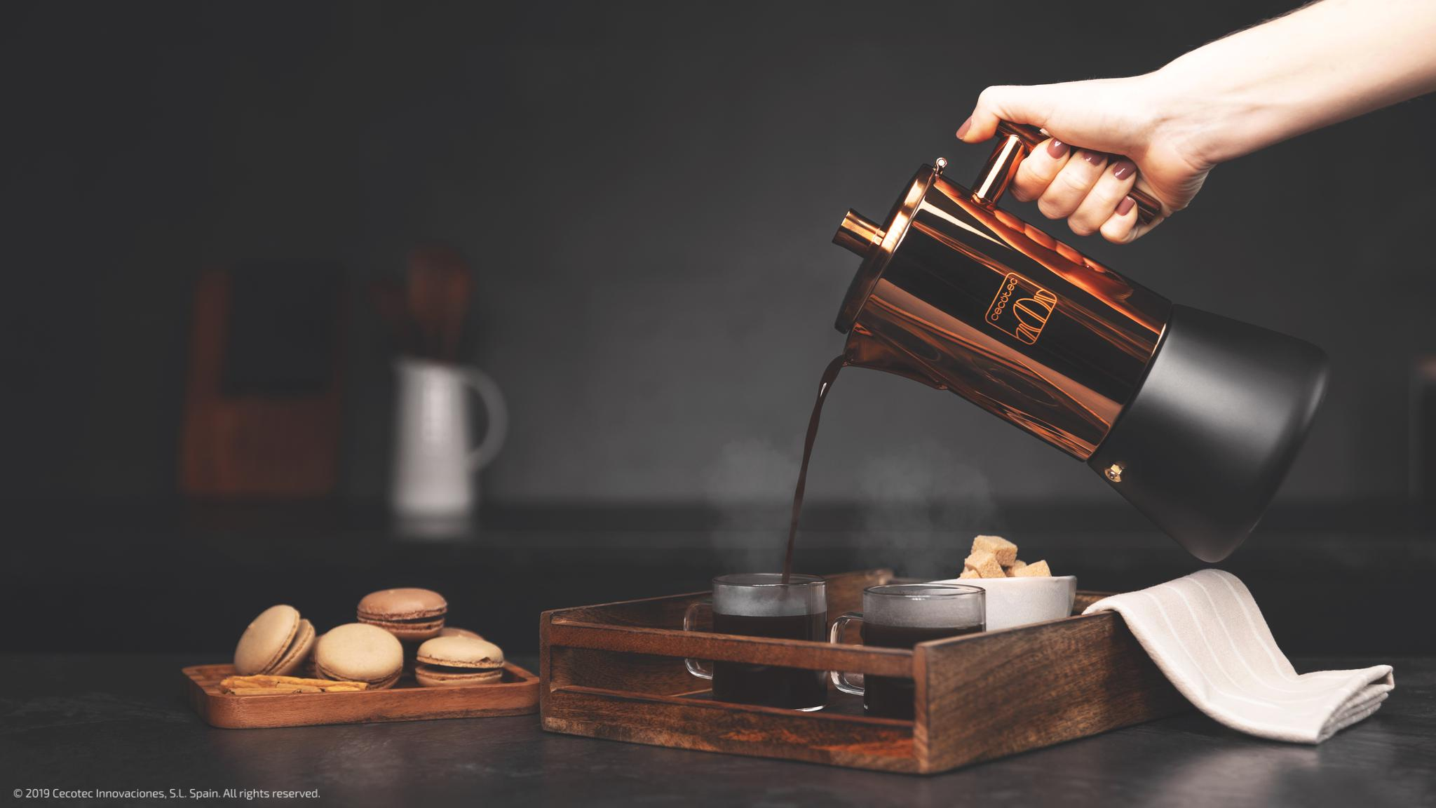 Una mano vierte café de una cafetera cobriza en tazas sobre una bandeja de madera con macarons cerca.