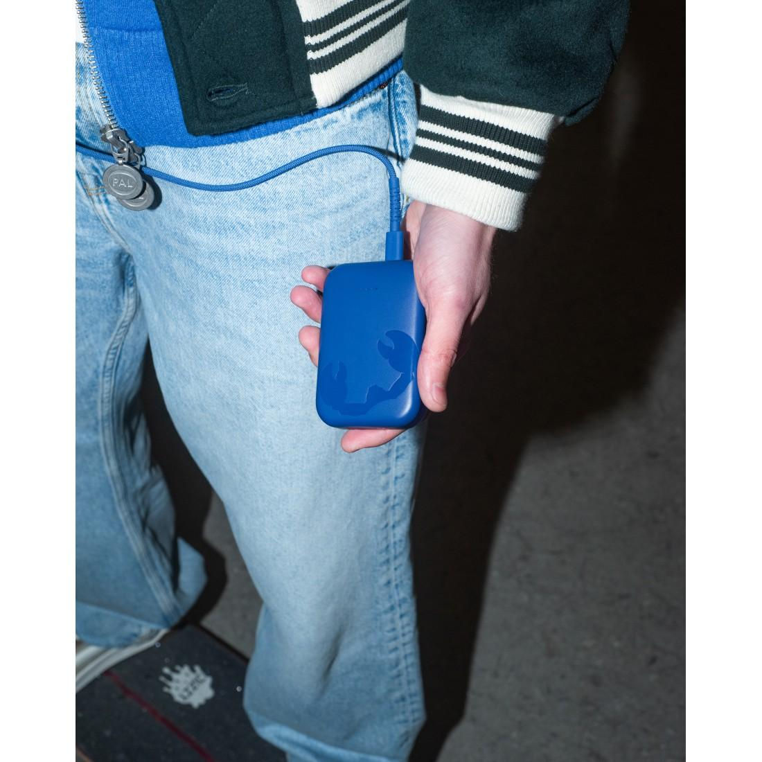 Person in Jeans und grüner Jacke hält ein blaues Gerät mit Kabel in der Hand, ein Skateboard steht auf dem Boden.