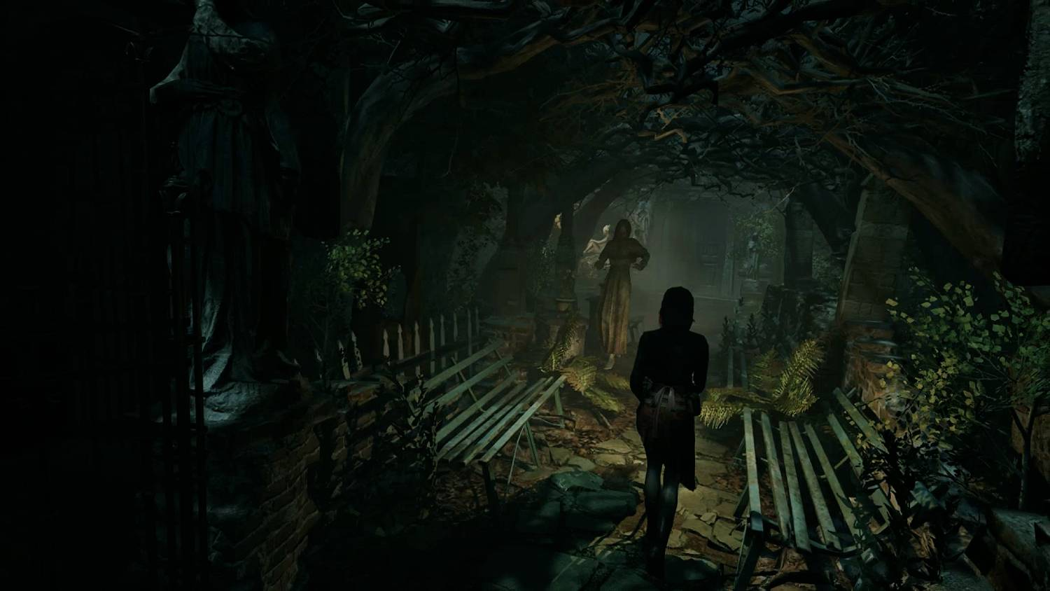 Una persona camina por un parque oscuro y cubierto de árboles con estatuas y bancos.