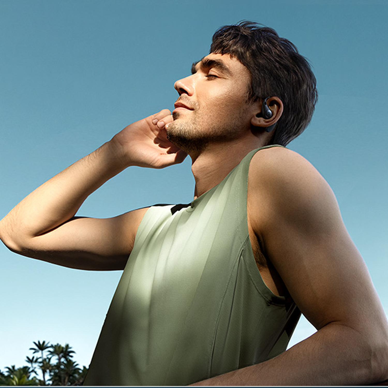 Hombre con los ojos cerrados, camiseta verde y auriculares disfruta del sol bajo un cielo azul.